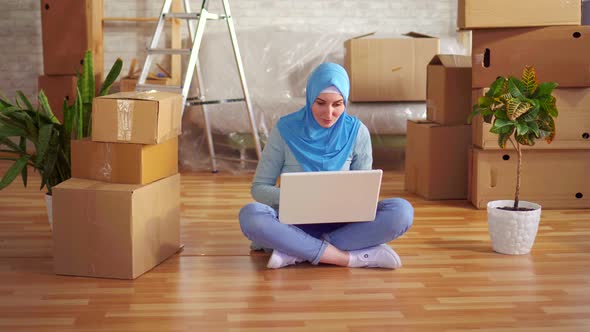 Young Muslim Woman in a Hijab Uses a Laptop Sitting on the Floor Next To the Boxes