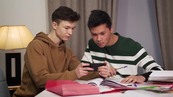 Two Modern Multiracial Friends Sitting at the Table and Using Smartphone. Smiling Teen College