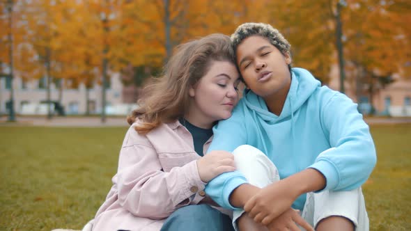 Diverse Young Women Sitting on Lawn in Autumn Park and Hugging