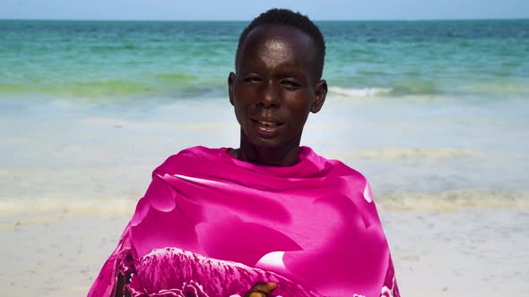 Native african man in pink clothing on beach, ocean waves behind.