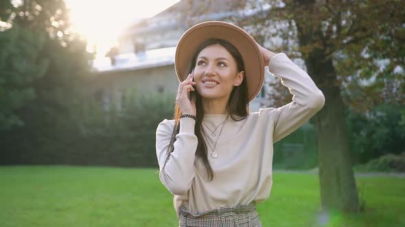 Young Woman in Beige Hat Standing Nearby Green Tree in the House's Yard and Talking on Phone