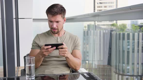 A Young Handsome Man Plays a Game on a Smartphone As He Sits on a Balcony in a Luxurious Apartment