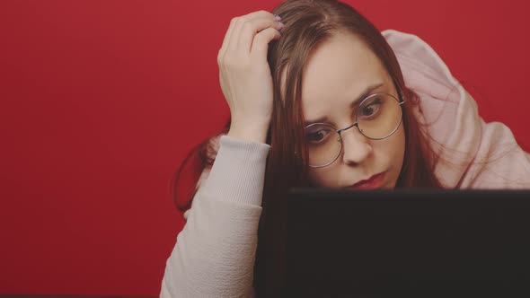 Young woman in glasses works on laptop and scratches head, sitting in home office.