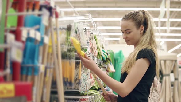 Slim Blonde Girl is Looking on a Garden Rake in a Supermarket Hall