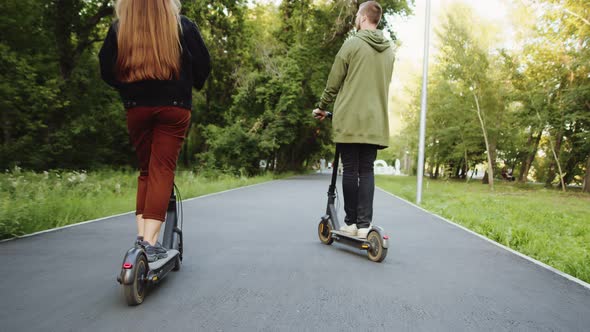 Man and Woman Ride Electric Scooters Along Alley in Public Park