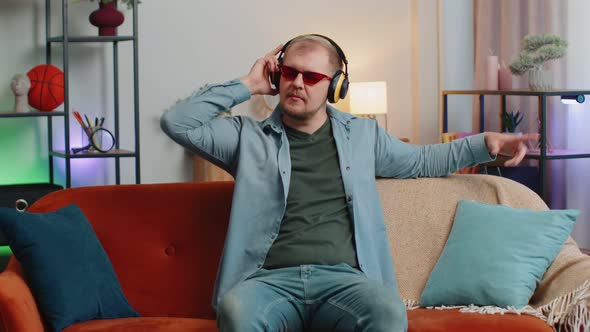 Overjoyed Young Man in Wireless Headphones Dancing Singing on Cozy Couch in Living Room at Home