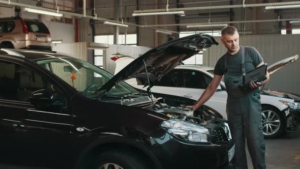 Handsome Mechanic in Uniform is Using a Laptop While Repairing Car in Auto