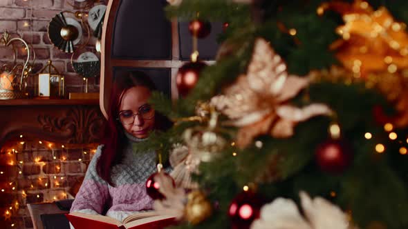 Concentrated Female in Warm Sweater and Eyeglasses Reading Book in Spare Time Near Christmas Tree