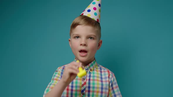 Elementary Age Boy Greeting with Happy Birthday Looking at Camera and Blowing Birthday Pipe on Blue