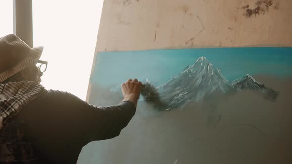 Old Artist Is Drawing By White Pastel Mountains on a Canvas