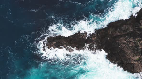 Aerial View of Waves Break on Rocks of Faroe Islands Cliffs in a Blue Ocean