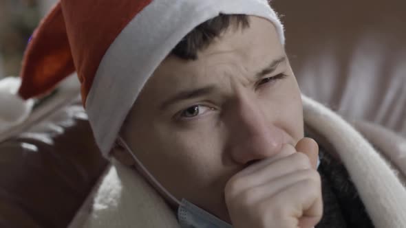 Closeup Headshot of Young Ill Man in Christmas Hat Coughing and Looking at Camera