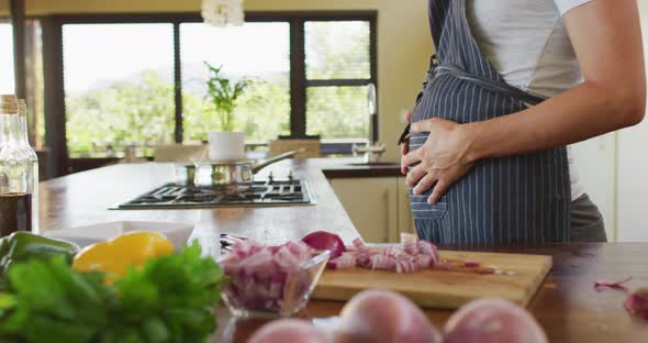 Midsection of caucasian pregnant woman wearing apron, touching belly in kitchen