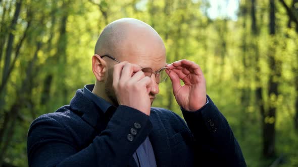 A Bald Stylish Man in a Jacket Puts on Glasses and Looks at the Camera