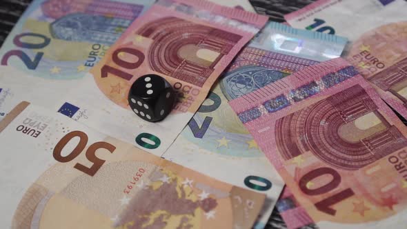Black dice rolls and rolls over on a table with euro banknotes.