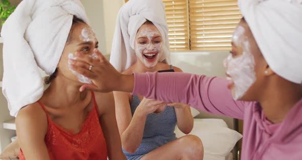 Diverse group of happy female friends with towels on heads and cleansing masks taking selfie at home