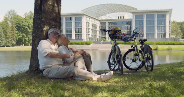 Senior Caucasian Couple Cyclists Sitting on Ground Under Tree Together in Park