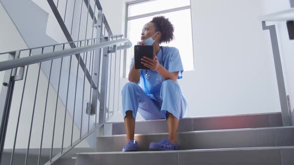 Mixed race female doctor wearing face mask sitting on stairs in hospital using tablet