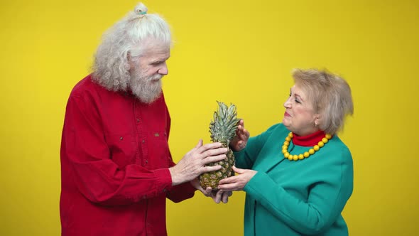 Positive Couple of Seniors Examining Pineapple and Talking on Yellow Background