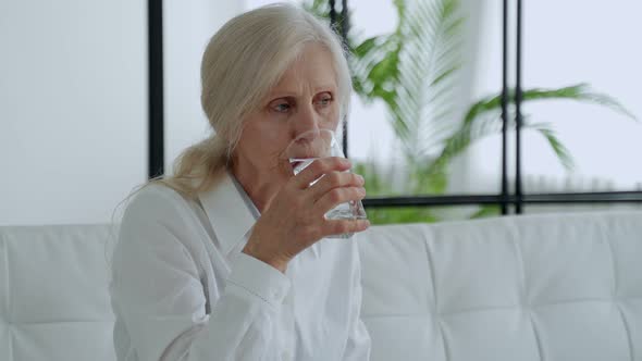 Elderly Woman Sits on a Sofa and Drinks Water From a Glass