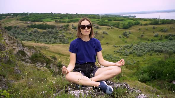Young Woman Mediating in Casual Clothes Sitting on a Rock in Nature Among Greenery