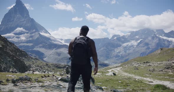 Black male traveler with backpack walking past screen towards cliff exploring the mountain landscape