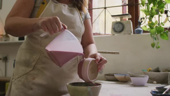 Mid section of female caucasian potter pouring paint from a jug on pot at pottery studio