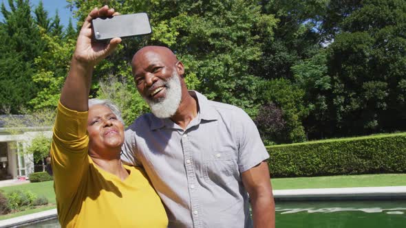 Happy senior african american couple using smartphone taking selfie by swimming pool in sunny garden