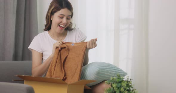 Happy Beautiful Asian Woman Opening Box With Ordered Clothes At Home On Couch.