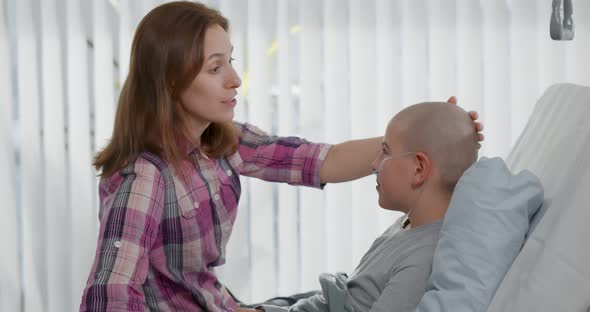 Side View of Woman Talking To Sick Son Resting in Bed After Chemotherapy in Hospital