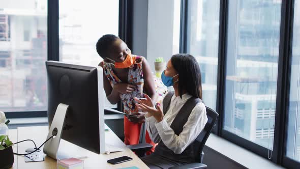 Diverse female office colleagues wearing face masks discussing at modern office