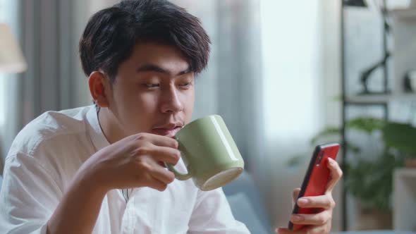 Close Up Side View Of Happy Asian Man Drinking Coffee While Using Smartphone In The Living Room