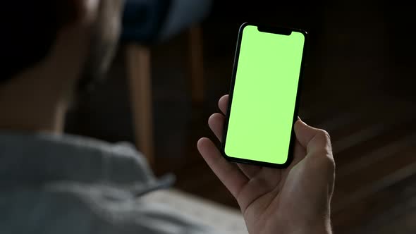 Man at home lying on a sofa and using smartphone with green mock-up screen in vertical mode