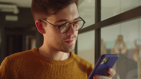 A Handsome Young Student Stands Near the Window Keeps a Phone in His Hands and Reads Something