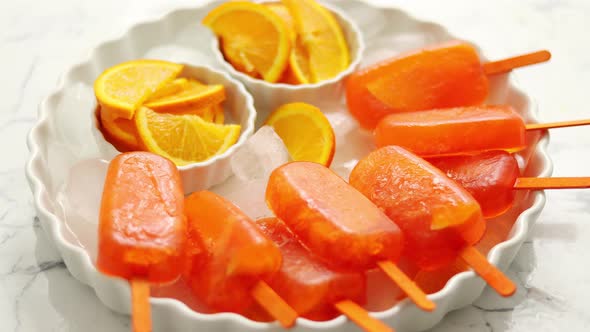 Homemade, Juicy, Orange Popsicles, Placed on a White Plate with Ice Cubes