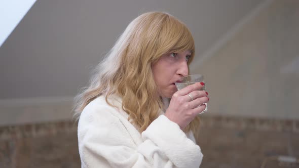 Side View Transgender Woman in Bathrobe Drinking Refreshing Water in Bathroom in the Morning