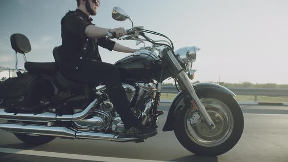 Biker rides motorcycle with chrome pipes on highway in countryside, side view. Man rides motorbike