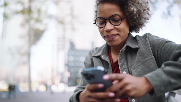 Happy adult African woman wearing casual clothes typing by mobile