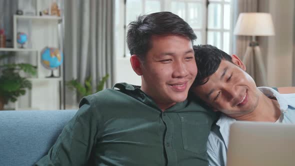 Asian Gentle Gay Couple Using Laptop Computer While Sitting On A Couch In Apartment