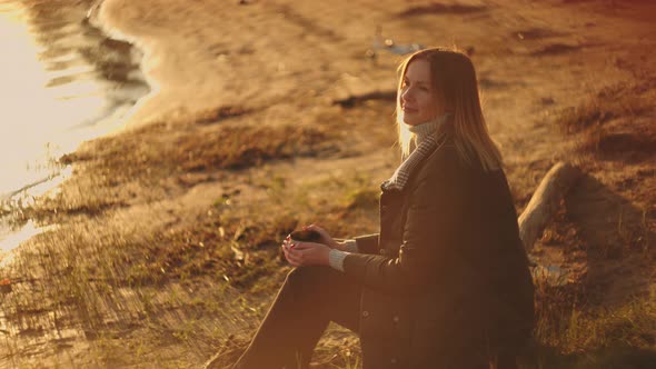 A Beautiful Young Woman Enjoys a Mug of Fragrant Drink in the Sun's Rays Watching the Sunset and on