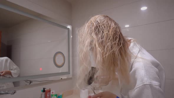 a blonde in white coat dries and styles her hair with hair dryer, in modern bathroom of house