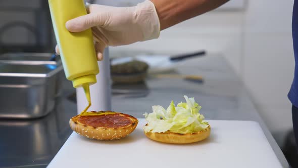 A hand in white gloves pouring sauce on toasted half of the bun. Making a delicious hamburger. 