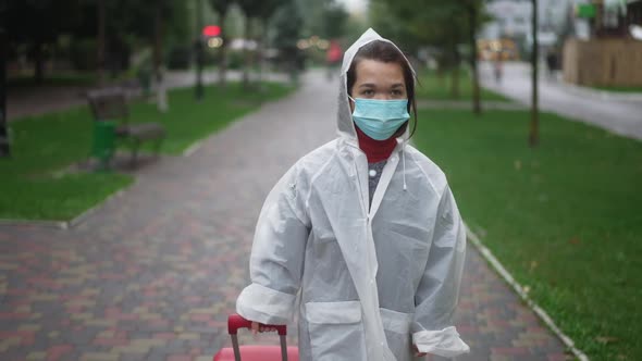 Dolly Shot of Young Little Person in Rain Coat and Coronavirus Face Mask Walking with Suitcase on