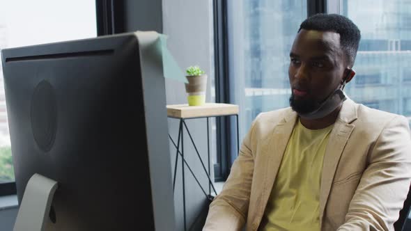 African american man with face mask around his neck using computer while sitting on his desk at mode