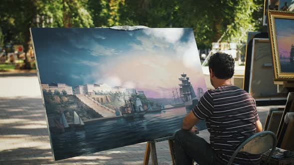 Man Artist Painting Marine Landscape with Sailing Ship and City on Coast While Sitting in Park