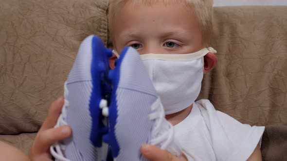 Sad Little Boy in a Mask Sitting on the Couch and Holding New Football Boots