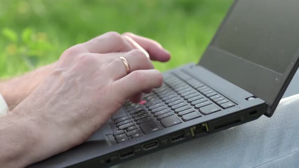 A Male User Uses His Hands to Type on a Laptop Keyboard While Sitting Outside in the Summer