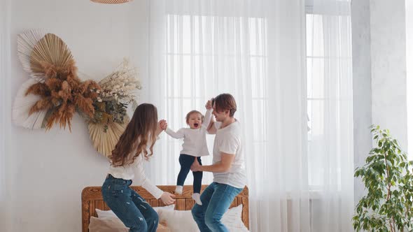 Young Parents Caucasian Mother and Millennial Father Standing on Bed at Home Raising Lifting Little