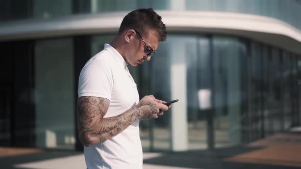Young Tattooed Man in Sunglasses Uses Smartphone, Business Center on Background