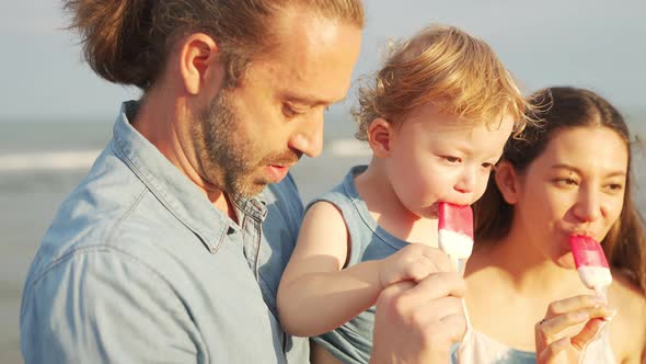 4K Caucasian parents with little baby son eating ice cream together at the beach.